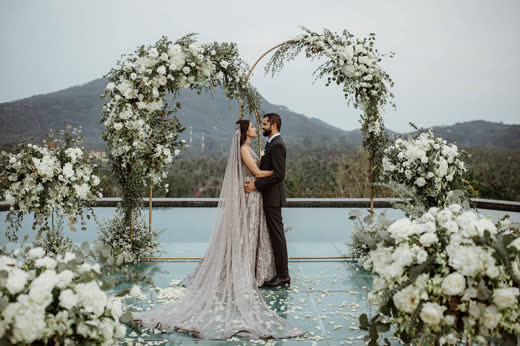 A couple under a white floral arch on a pool edge with hills behind